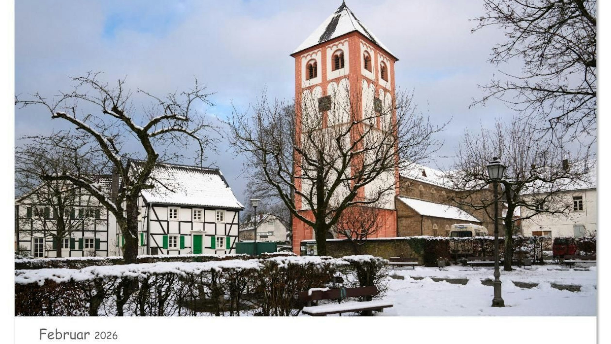 Ein Kirchturm bedeckt mit Schnee steht hinter kahlen Bäumen.