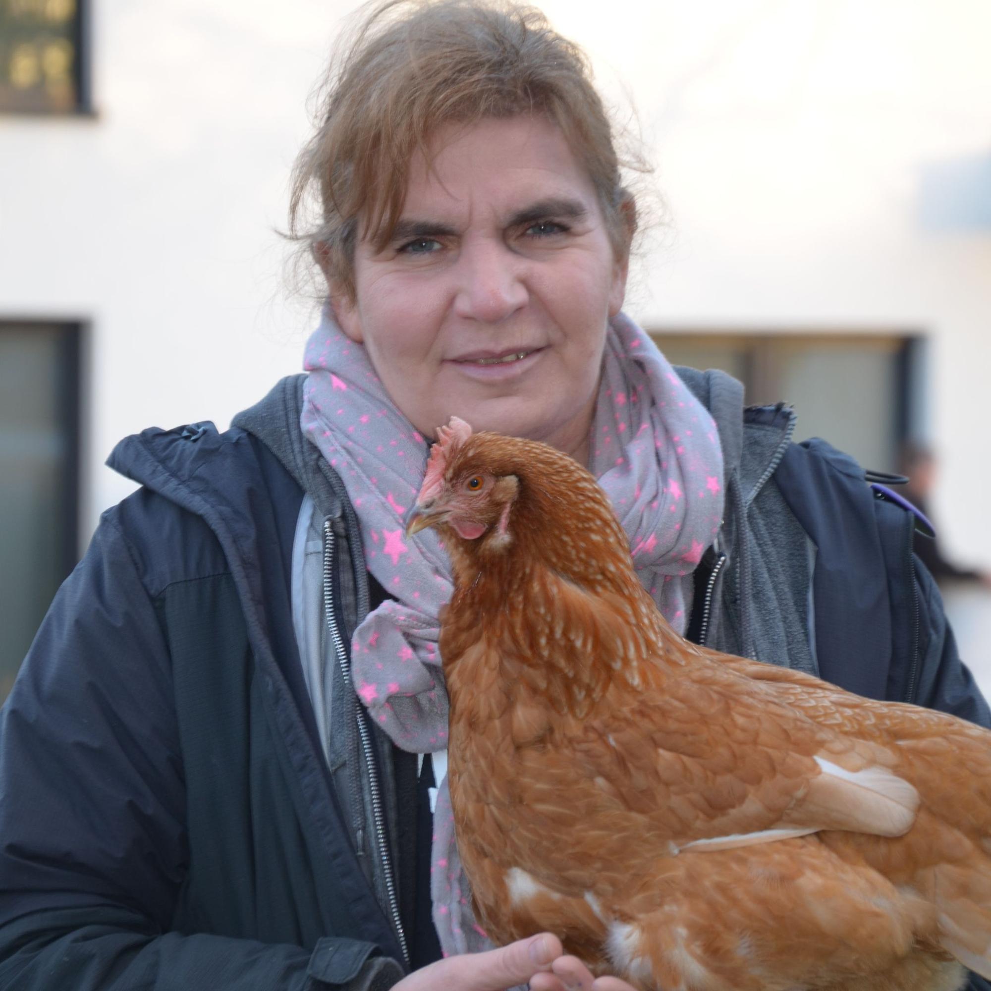 Auf dem Bild ist eine Frau zu sehen, die ein braunes Huhn auf dem Arm hält.