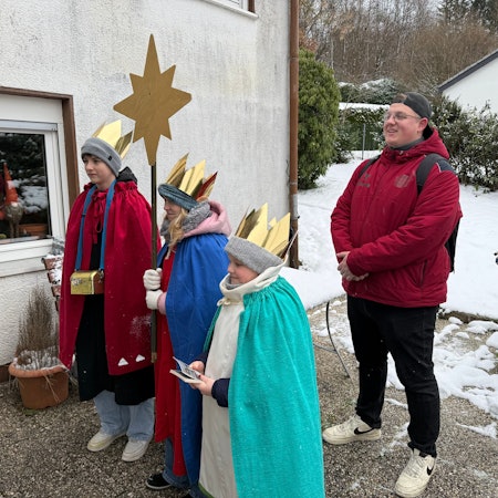 Drei Kinder stehen als Sternsinger vor einer Tür. Dahin steht ihr Begleiter