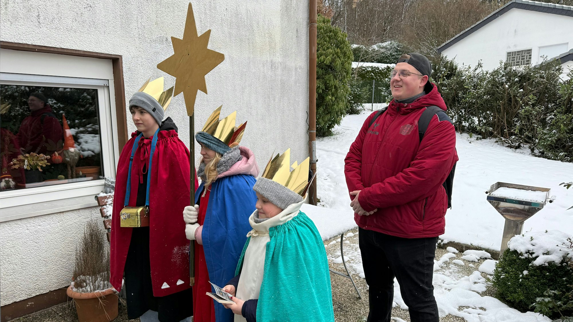 Drei Kinder stehen als Sternsinger vor einer Tür. Dahin steht ihr Begleiter