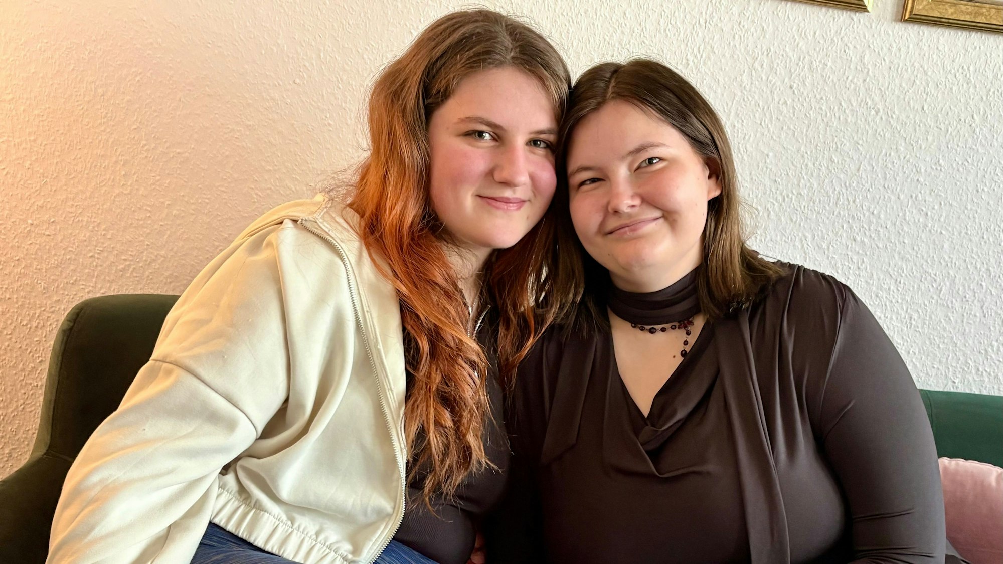 Die beiden jungen Frauen sitzen eng aneinander gekuschelt auf einem Sofa und lächeln.