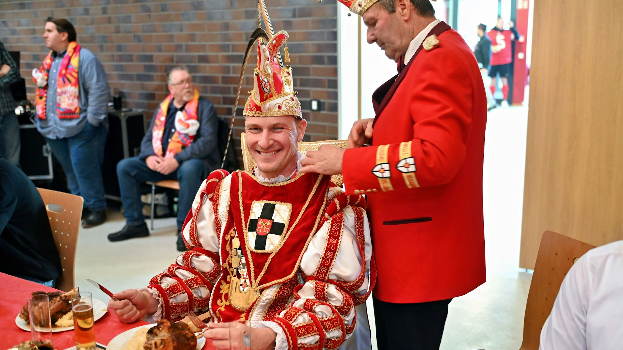 Ein Mann im Prinzenornat bekommt ein Lätzchen umgebunden. Vor ihm steht ein Teller mit einer Haxe.