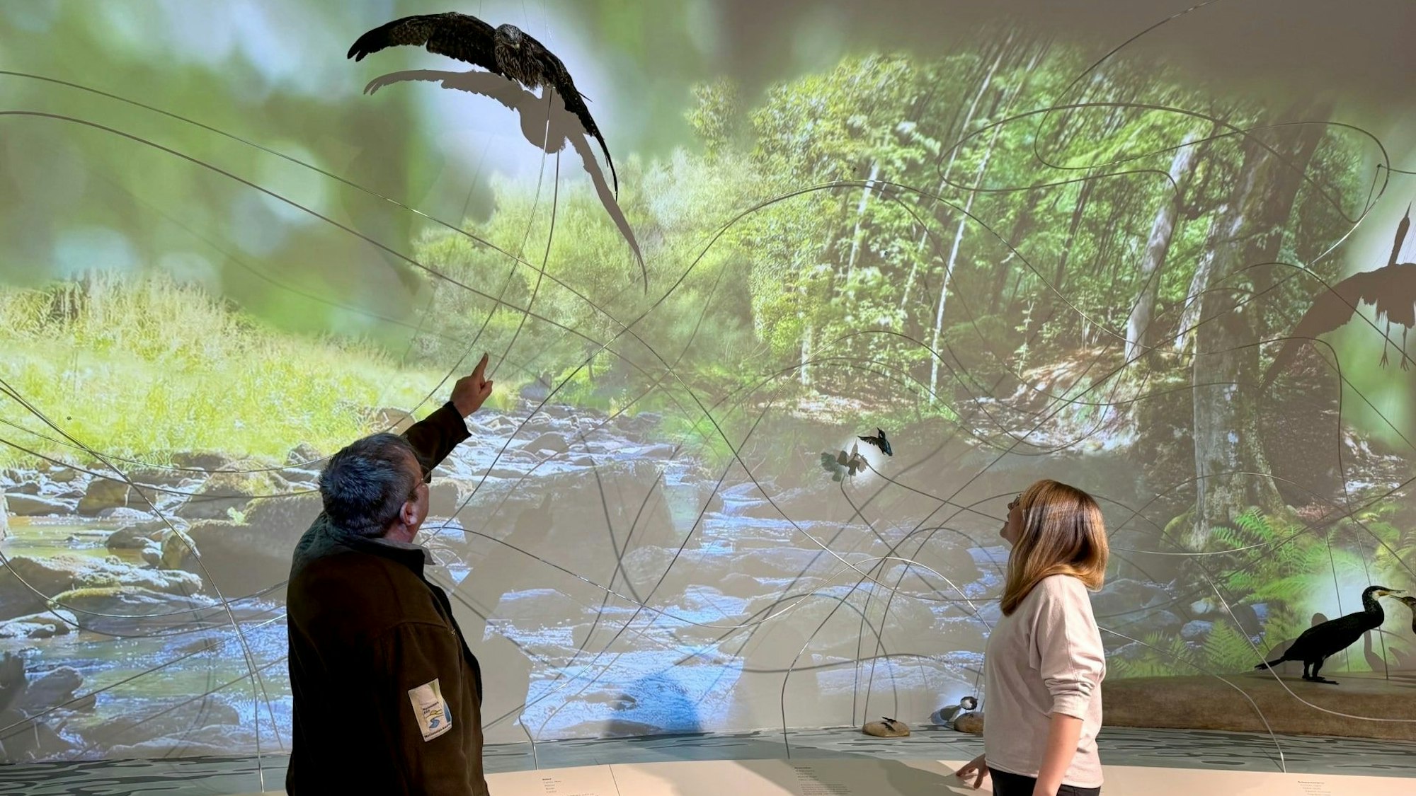 Ein Mann und eine Frau stehen vor einer Leinwand, auf der eine Waldlandschaft mit einem Bach zu sehen ist.