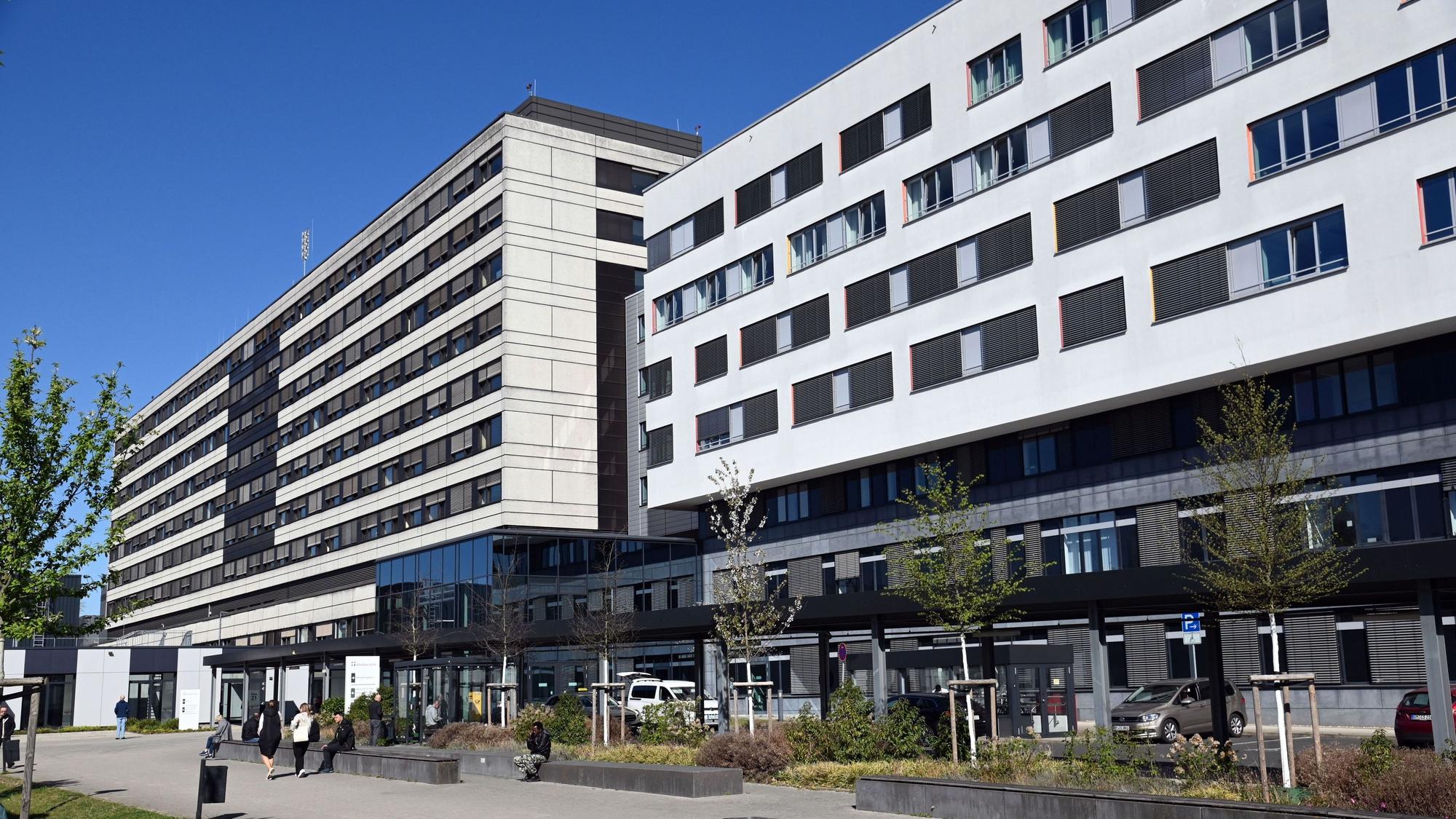 Ein großer Klinikkomplex mit mehrstöckigen Gebäuden, Vordach und Vorplatz, auf dem Menschen sitzen und gehen, unter klarem blauem Himmel.