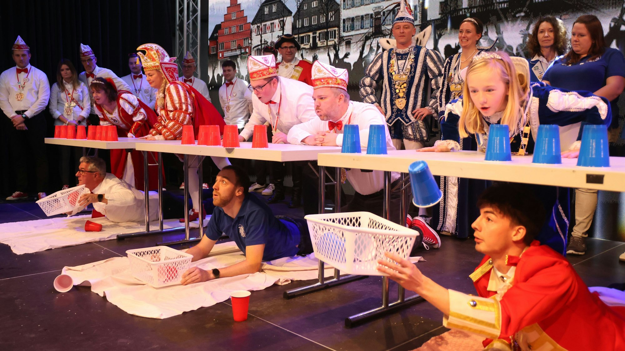 Das Bild zeigt ein kurzweiliges Spiel der Narren: Die einen pusten Plastikbecker über die Tischkante, die anderen liegen bäuchlings unterm Tisch und fangen die Becher mit einem Körbchen auf.