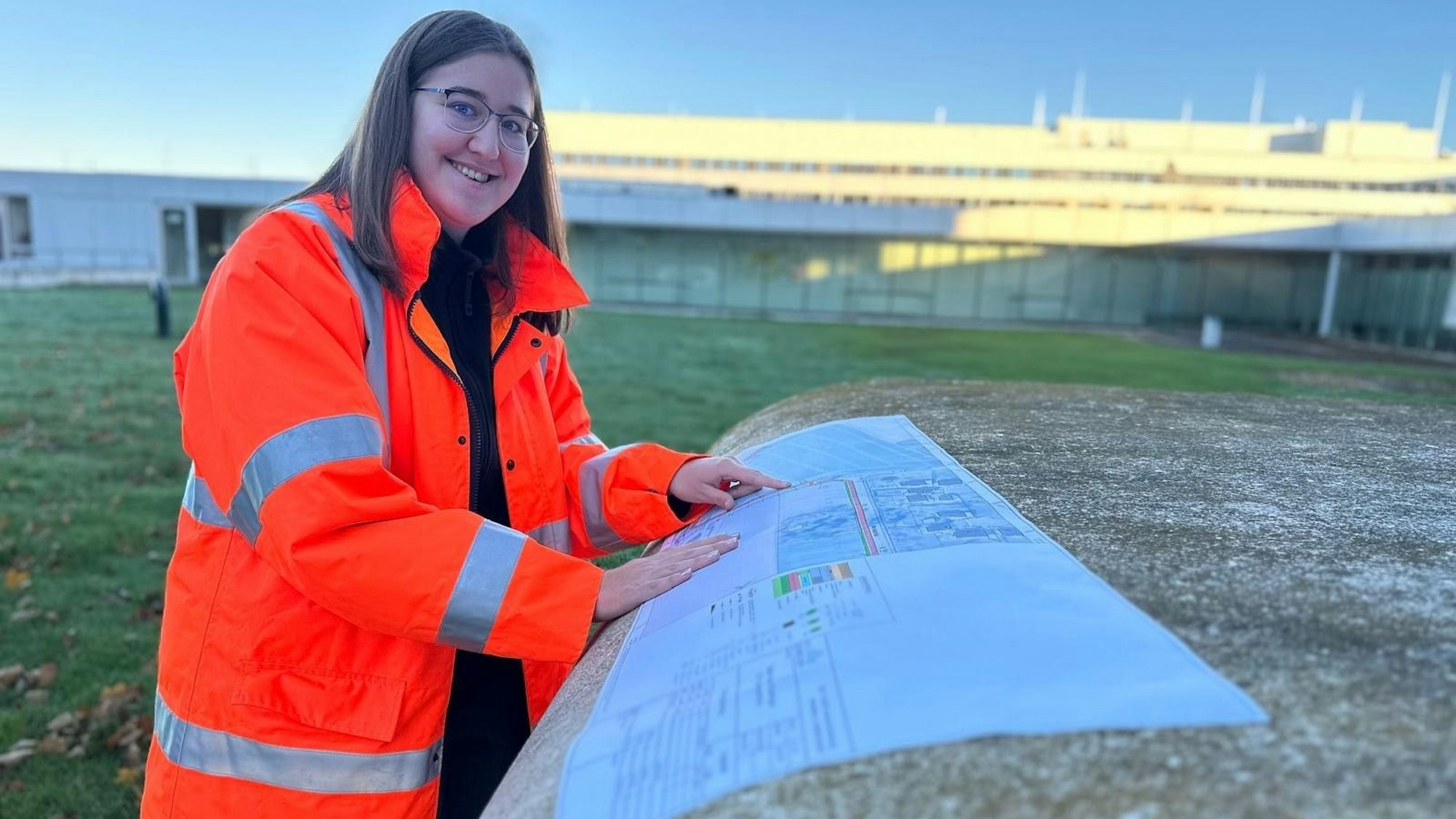 Hannah Bär trägt eine Warnweste und hat ihre Hände auf einen Bauplan gelegt. Sie steht draußen. Im Hintergrund fällt die Sonne auf einen unscharf gestellten Gebäudekomplex.