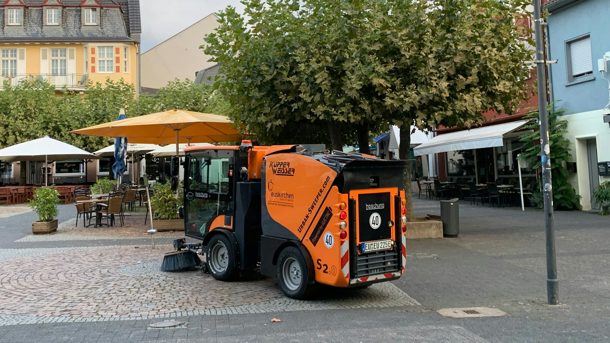 Eine Kehrmaschine reinigt den Platz, im Hintergrund stehen Sonnenschirme.