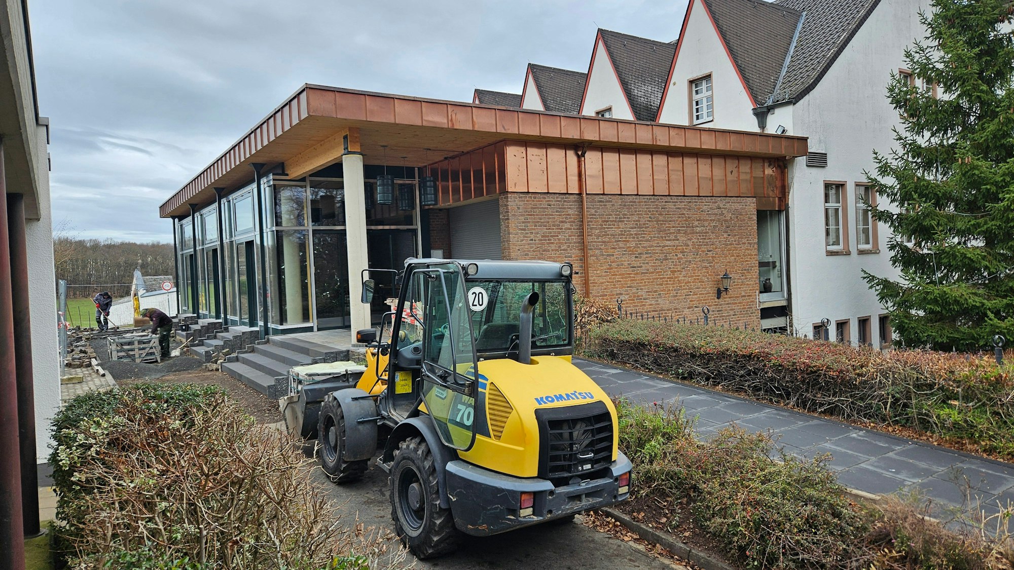 Zwei Arbeiter verlegen im Hintergrund das Pflaster. Im Vordergrund ist ein Radlader zu sehen.