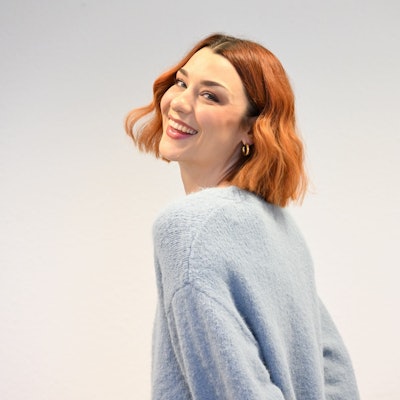 Eine junge Frau mit rotem Haar lächelt über ihre Schulter hinweg in die Kamera.