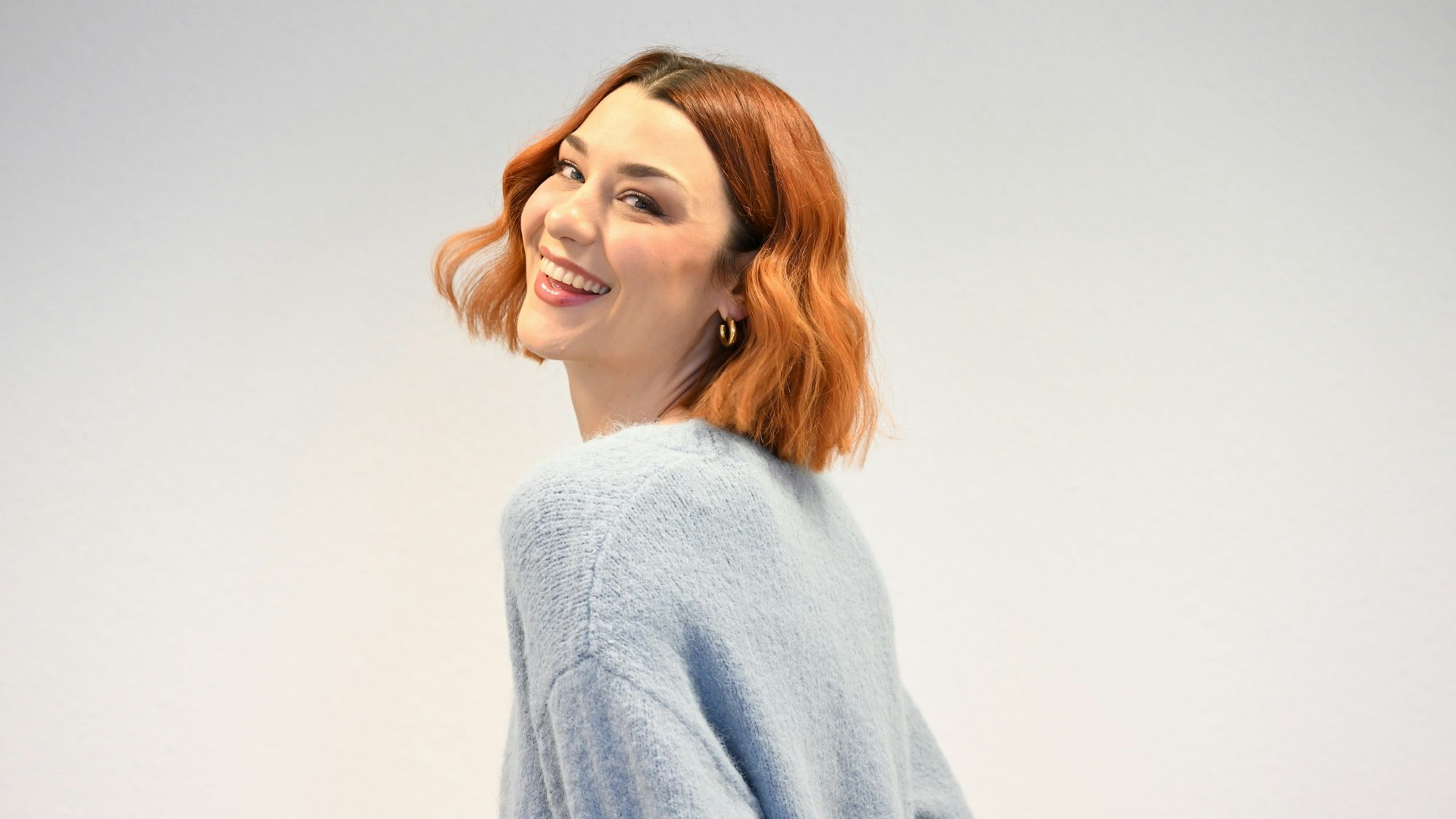 Eine junge Frau mit rotem Haar lächelt über ihre Schulter hinweg in die Kamera.