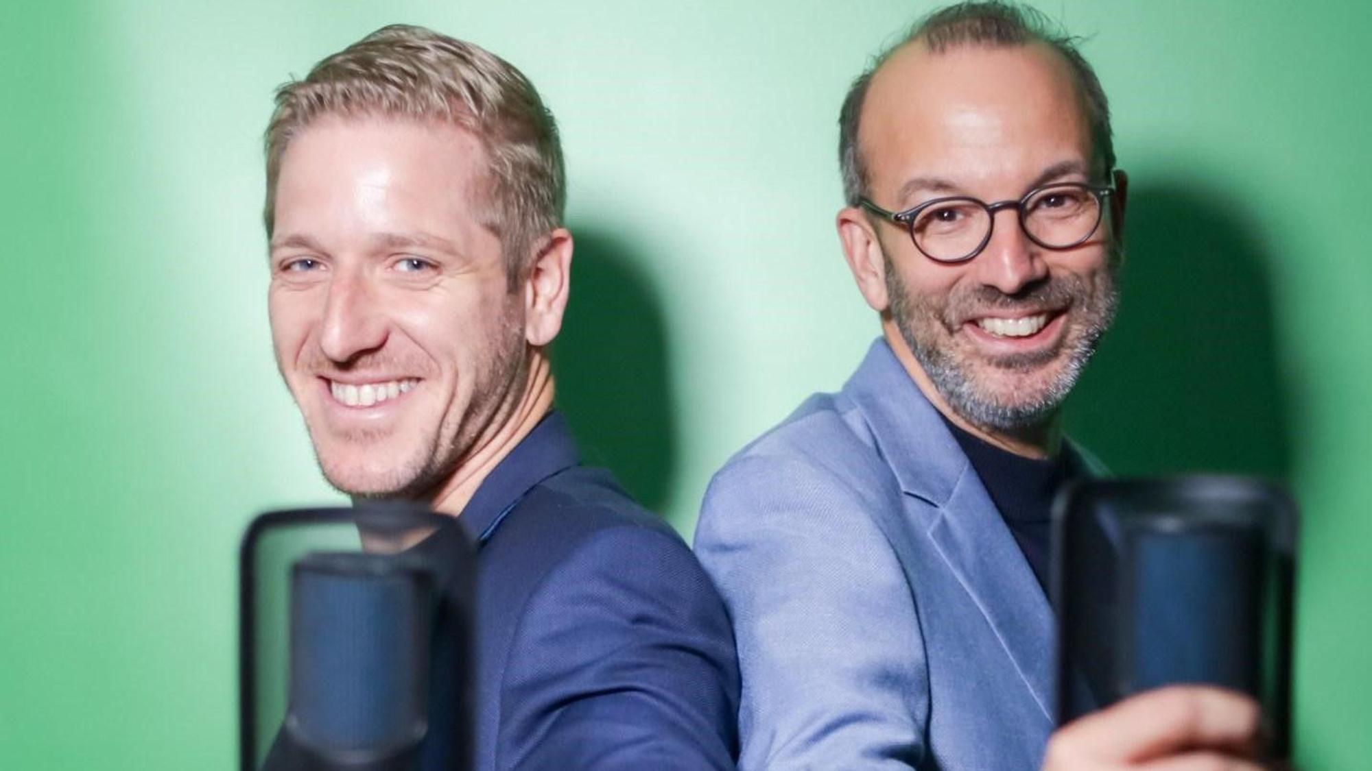 Das Bild zeigt die beiden Obengenannten, die für ein Foto posieren. In der Hand halten sie jeweils ein Mikrofon.