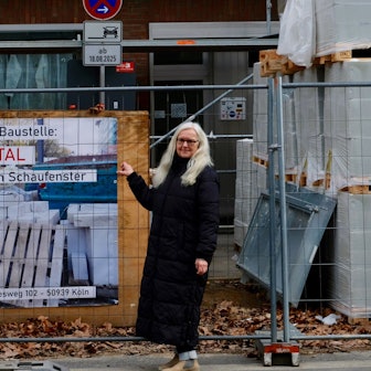 Eine Frau im schwarzen Mantel mit langem blonden Haar steht an einem Baustellenzaun. Dahinter prangt ein Plakat mit der Aufschrift Frontal-Kunst im Schaufenster.