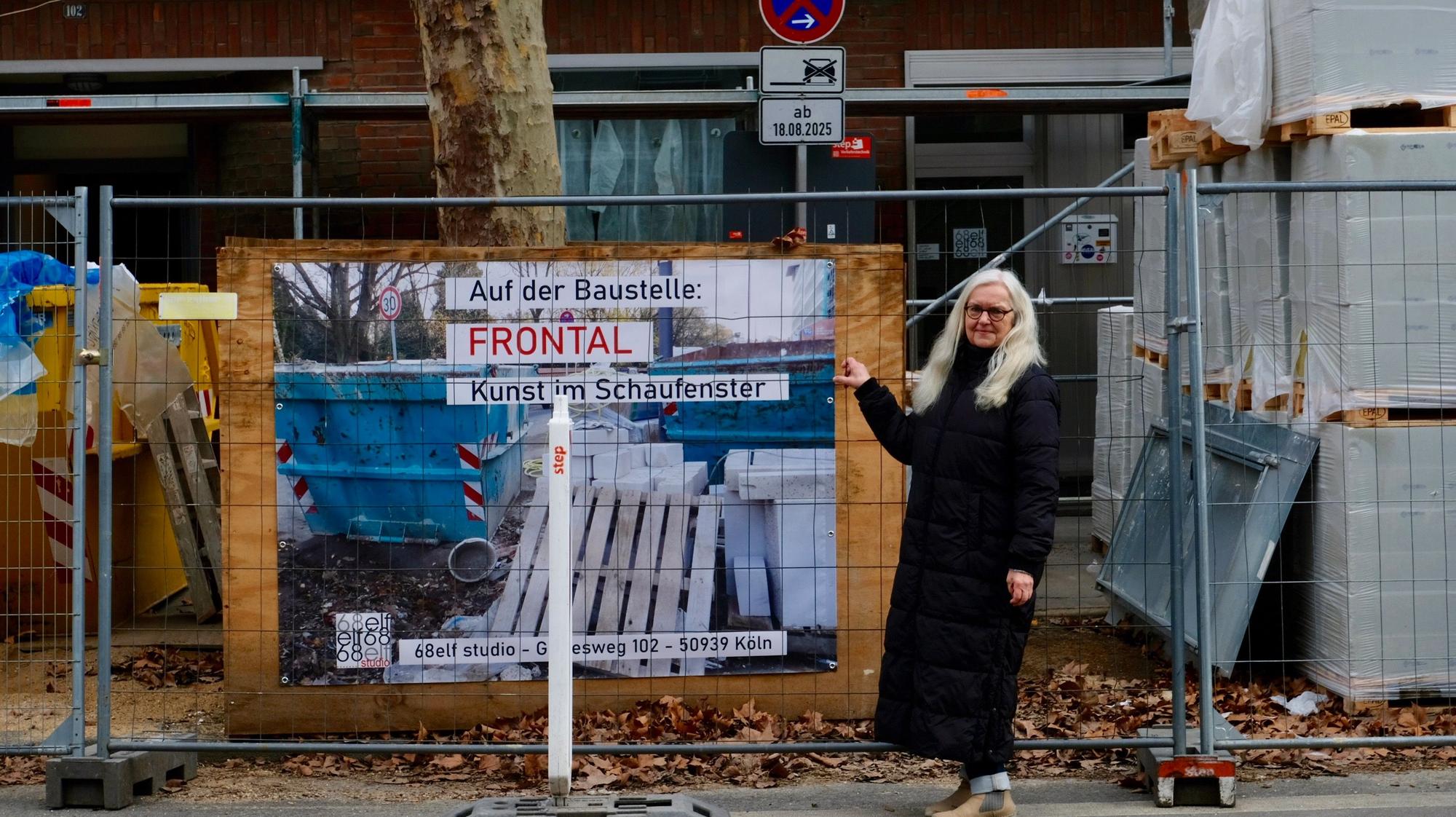 Eine Frau im schwarzen Mantel mit langem blonden Haar steht an einem Baustellenzaun. Dahinter prangt ein Plakat mit der Aufschrift Frontal-Kunst im Schaufenster.