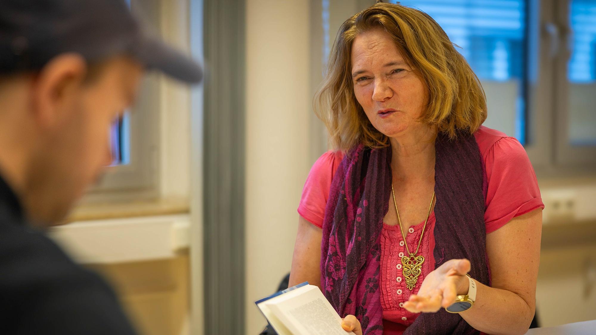 Gunild Lohmann-Sistig sitzt und hält mit der rechten Hand ein aufgeschlagenes Buch, während sie mit der linken Hand gestikuliert. Im Vordergrund befindet sich ein unscharf gestellter Mann mit Kappe.