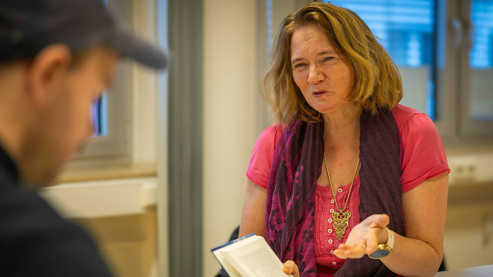 Gunild Lohmann-Sistig sitzt und hält mit der rechten Hand ein aufgeschlagenes Buch, während sie mit der linken Hand gestikuliert. Im Vordergrund befindet sich ein unscharf gestellter Mann mit Kappe.