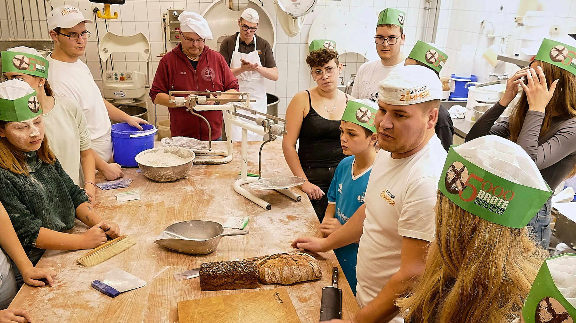 Kinder und Jugendliche stehen an einem Tisch mit Backutensilien, ein Bäcker ist rechts im Bild zu sehen.