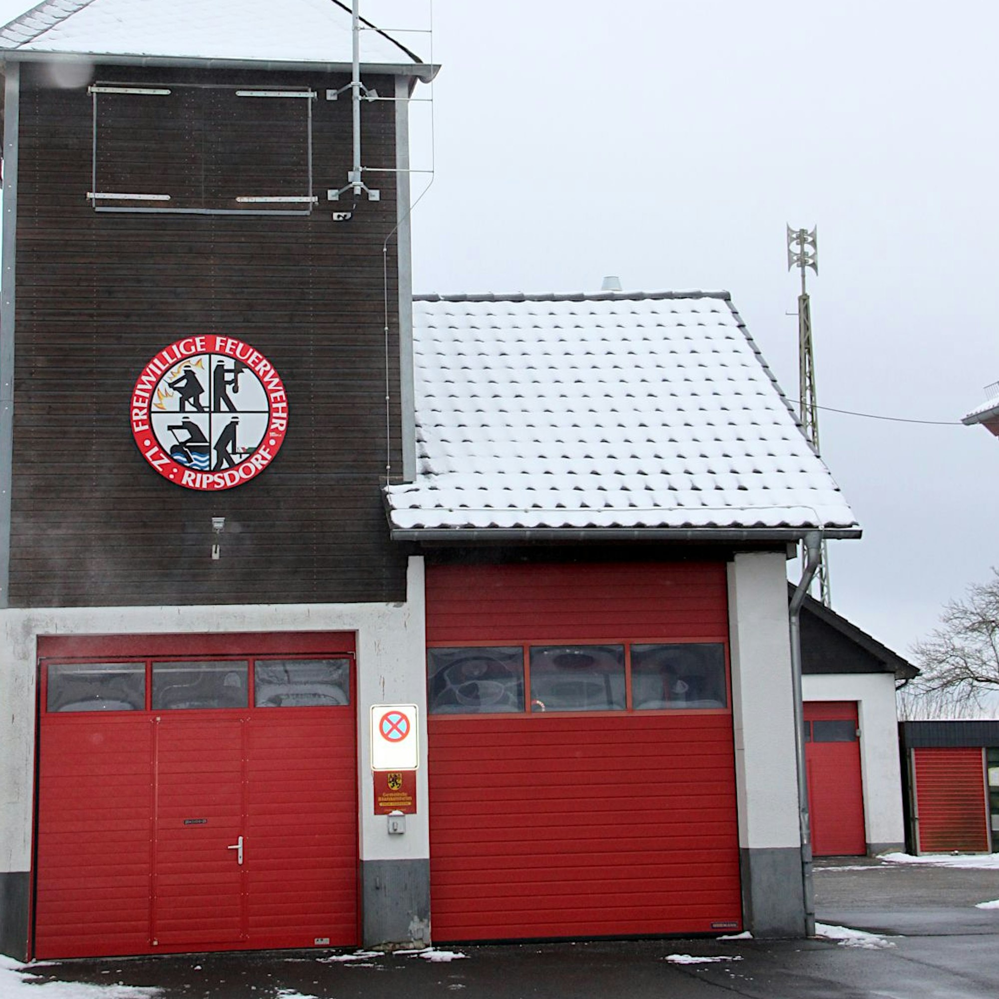 Das Gerätehaus hat zwei rote Rolltore und das Feuerwehr-Emblem an der Wand des Turms.