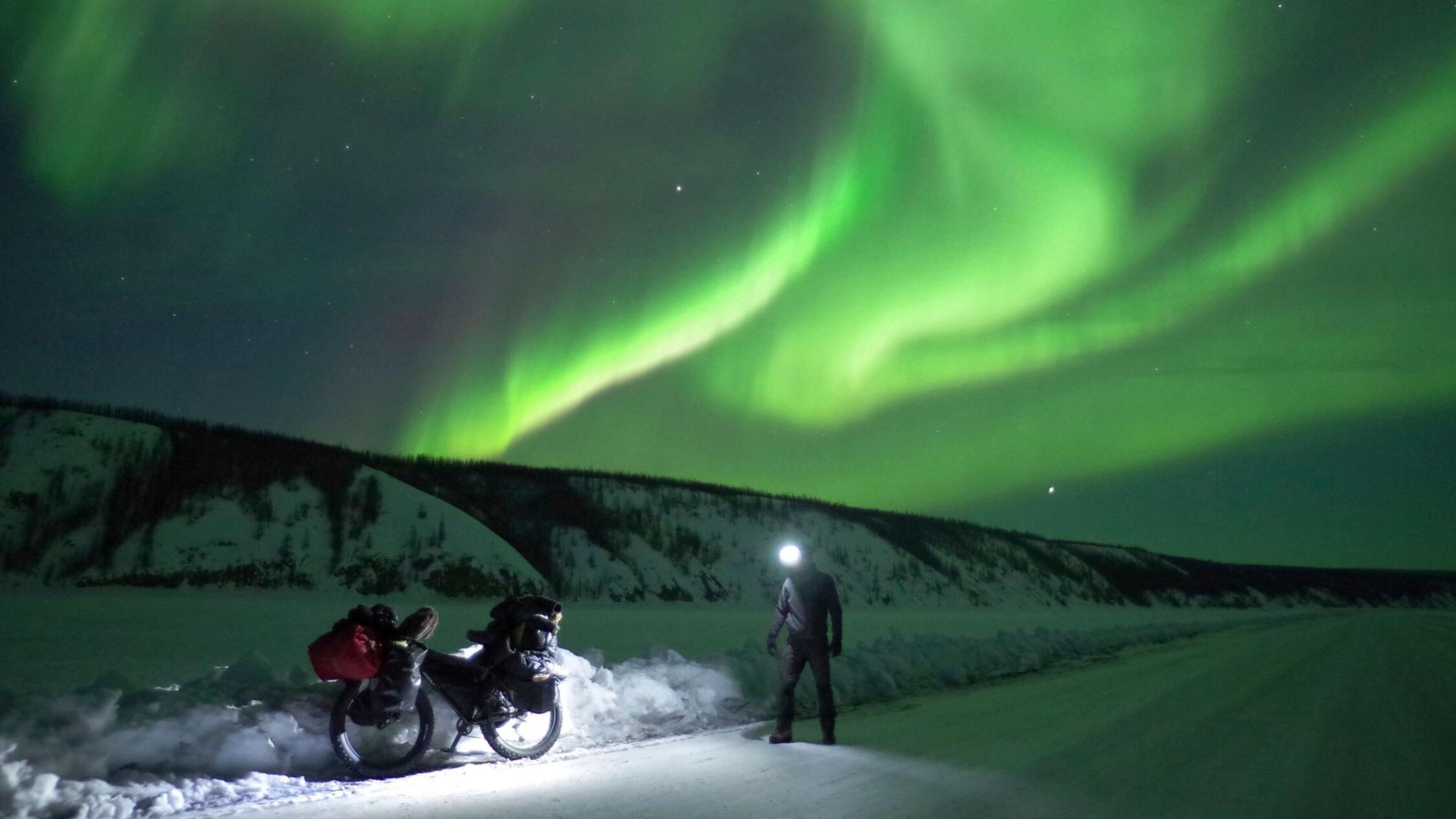 Das Bild zeigt grün schimmernde Polarlichter am Himmel und einen Radfahrer mit Stirnlampe im Vordergrund.
