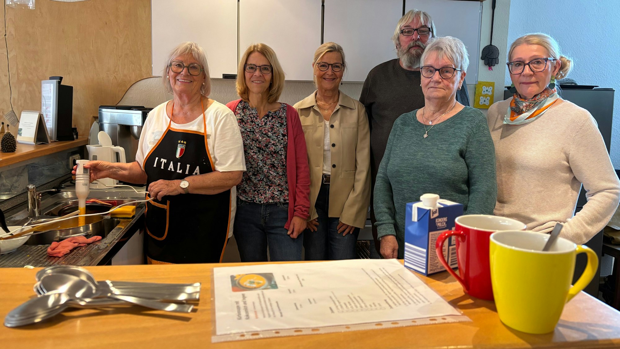 Fünf Frauen und ein Mann, die allesamt Brillen tragen, stehen in einer Küche. Im Vordergrund befinden sich Kaffeetassen und Löffel auf einer Theke.
