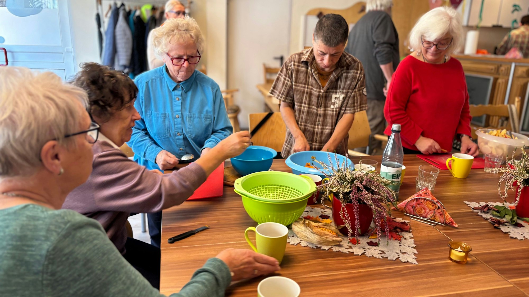 Vier ältere Frauen und ein Mann bereiten an einem Tisch eine Suppe zu. Auf dem Tisch liegen Schalen für geschnittene Zutaten und Schneidebretter.