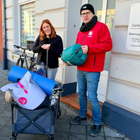Die beiden Sozialarbeiter der Caritas stehen vor dem Gebäude der Wohnungslosenhilfe. Sie haben einen blauen Bollerwagen dabei, auf dem sich Decken, Schlafsäcke und Isomatten befinden.