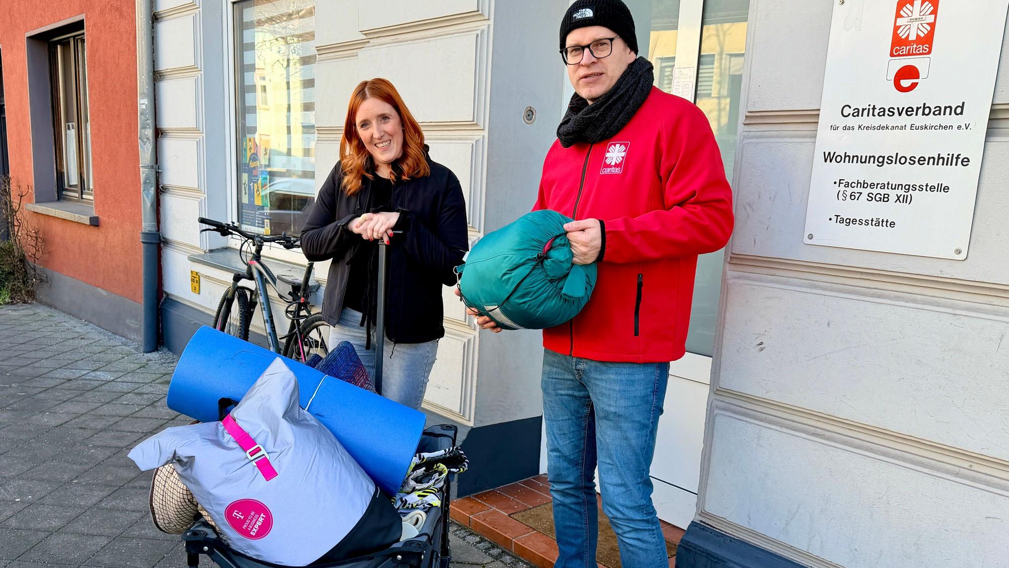 Die beiden Sozialarbeiter der Caritas stehen vor dem Gebäude der Wohnungslosenhilfe. Sie haben einen blauen Bollerwagen dabei, auf dem sich Decken, Schlafsäcke und Isomatten befinden.