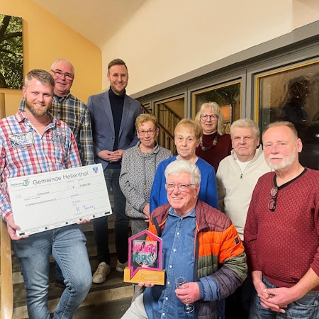 Neun Menschen haben sich zum Gruppenbild versammelt. Ein Mann hält einen überdimensionalen Scheck in den Händen, ein anderer einen Preis.