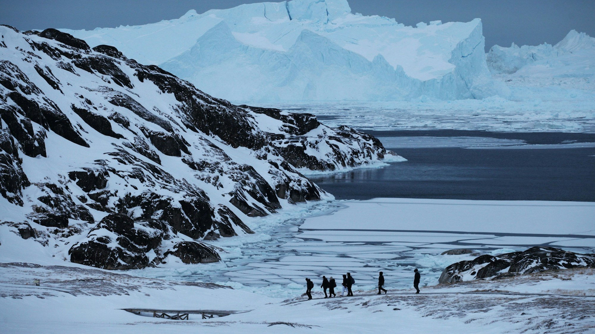 Eine Gruppe Touristen wandert in der Nähe des grönländischen Ortes Ilulissat vor der Kulisse riesiger Eisberge entlang.