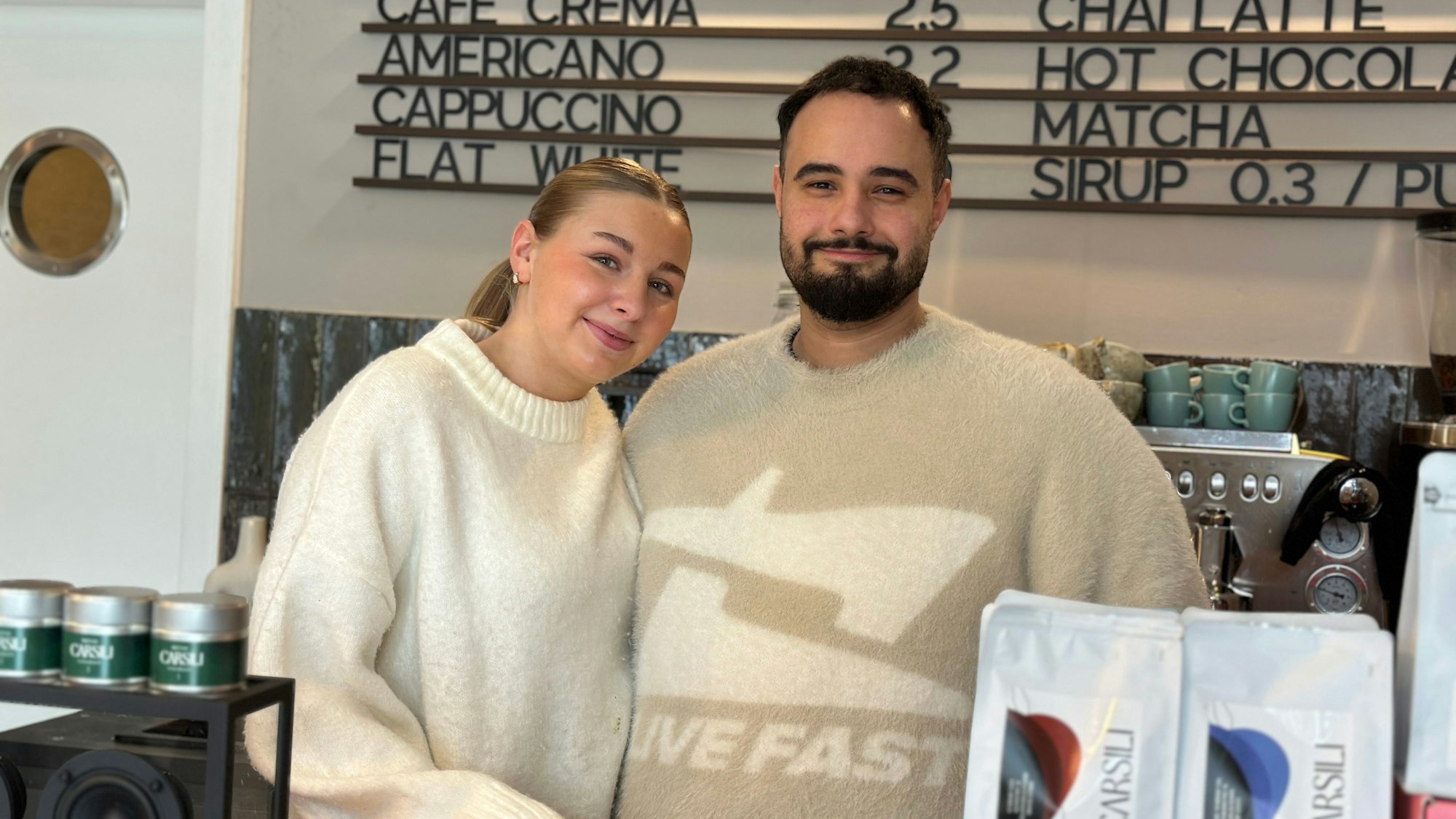 Eine blonde Frau und ein Mann mit dunklem Haar und Bart stehen hinter dem Verkaufstresen eines Cafés.