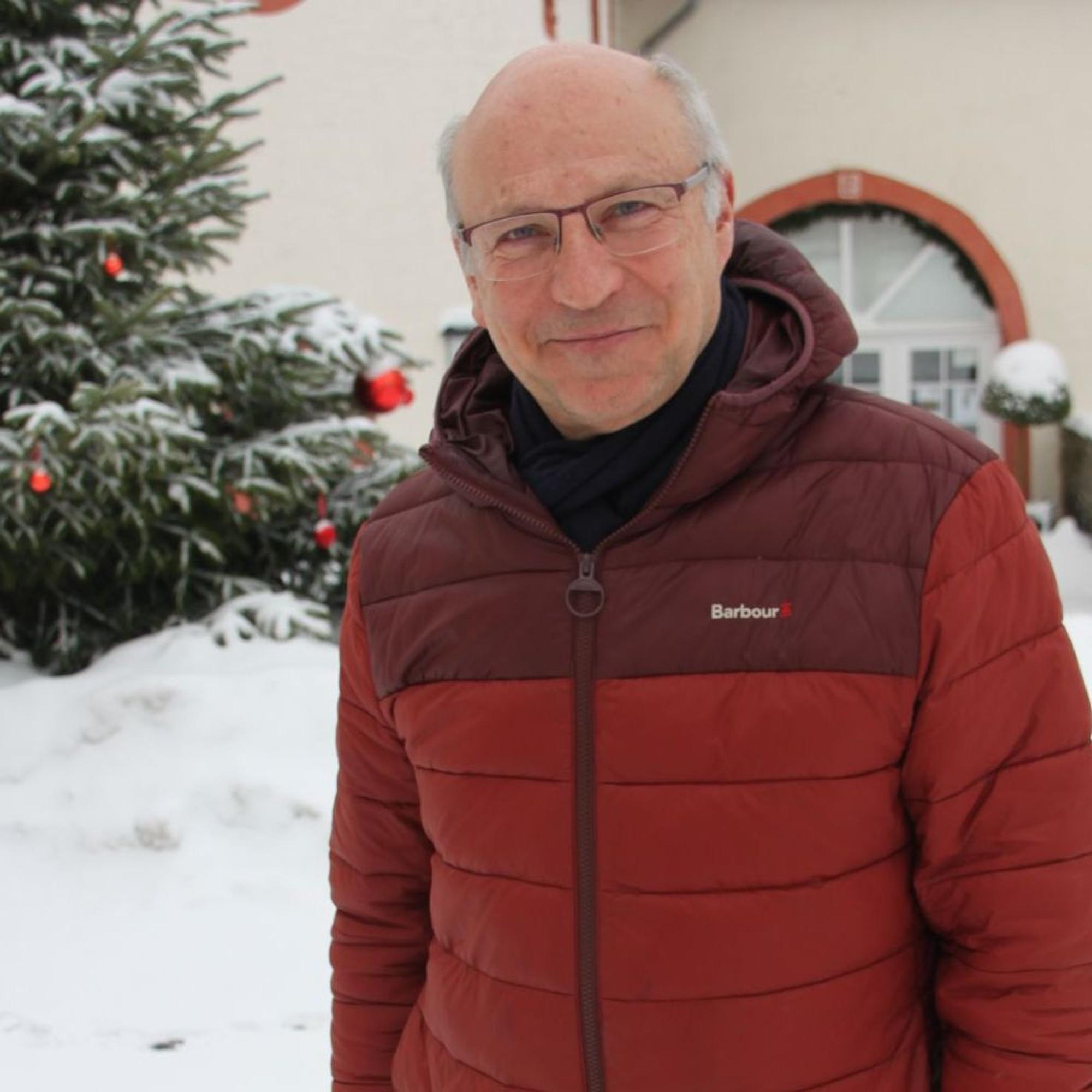 Ein Mann, der eine rote Winterjacke trägt, steht im Schnee vor einem historischen Gebäude und einem Weihnachtsbaum.