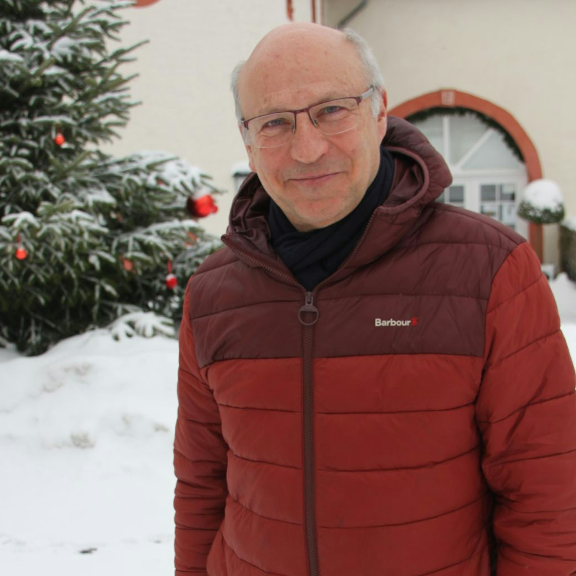 Ein Mann, der eine rote Winterjacke trägt, steht im Schnee vor einem historischen Gebäude und einem Weihnachtsbaum.