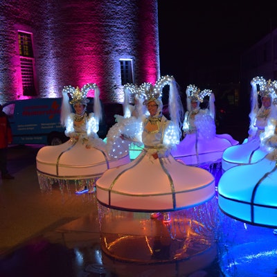 Frauen in weit ausfallenden Kostümen, die an pompöse Kleider erinnern, stehen auf einer glatten Fläche, in der sich die leuchtenden Anziehsachen spiegeln.