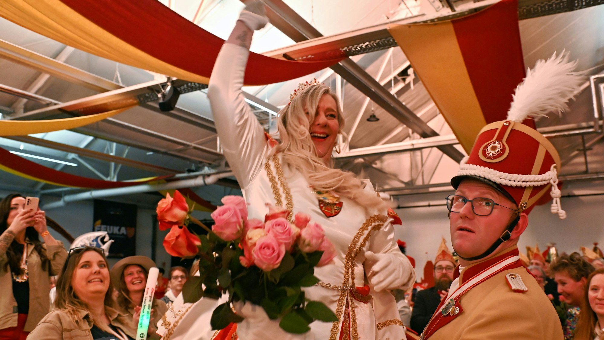 Prinzessin Colette I. in einem weißen Prinzenkleid beim Einzug in den Saal. Rechts steht ein Adjutant, der ihr einen Strauß Rosen anreicht.