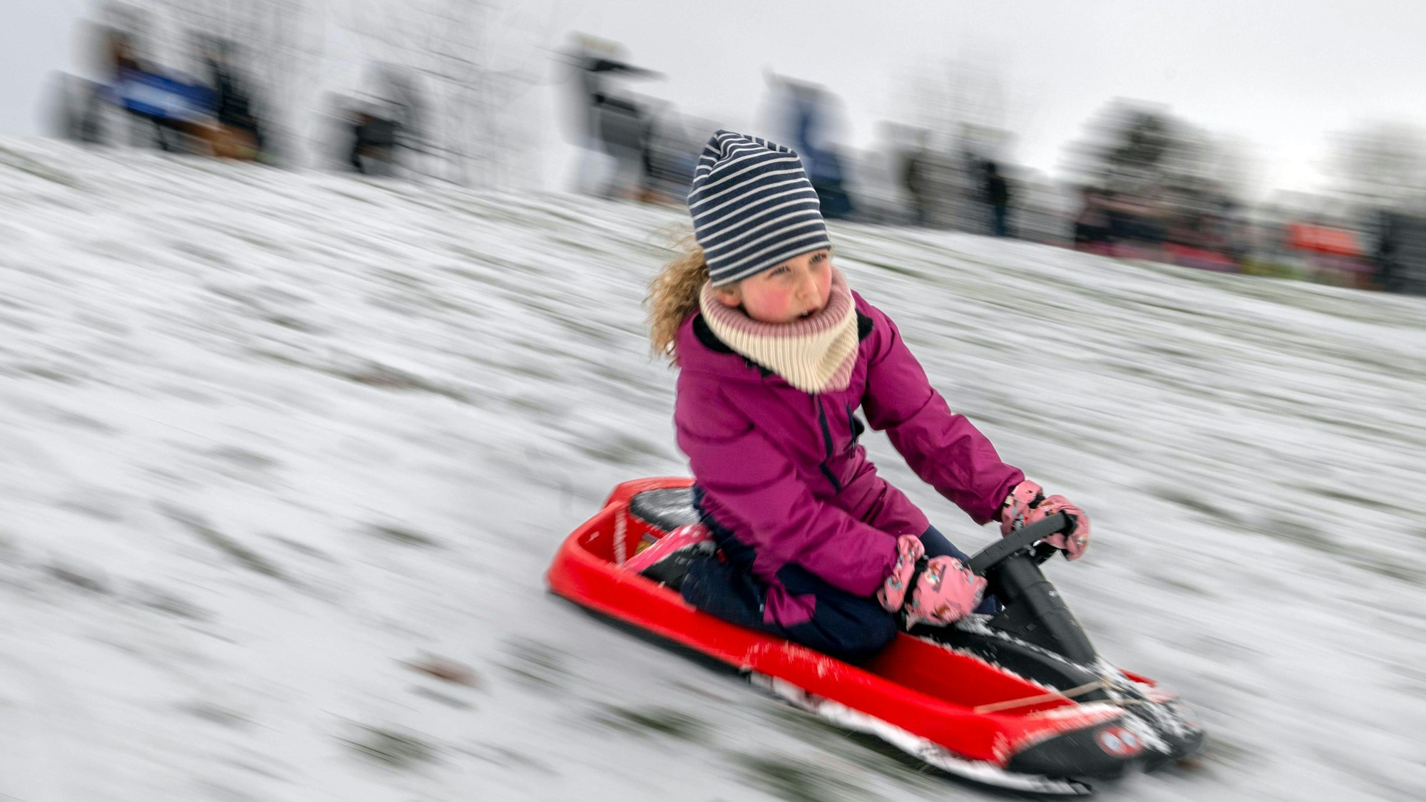 Ein Mädchen mit Mütze, Schal und lilafarbener Winterjacke rodelt auf einem roten Schlitten einen Hang hinunter.