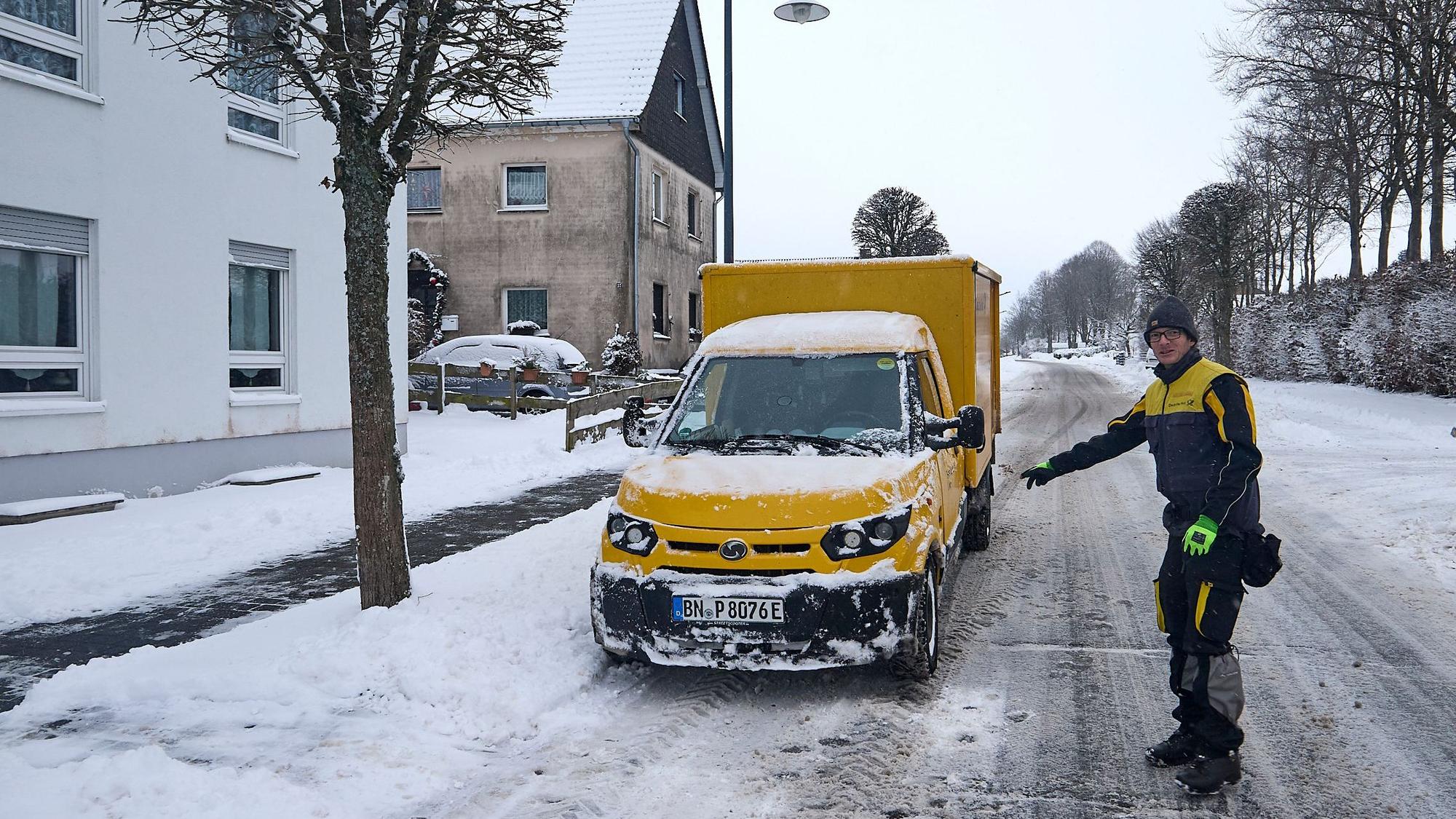 Ein Zusteller der Post geht über eine schneebedeckte Straße zu seinem elektrischen Zustellfahrzeug.