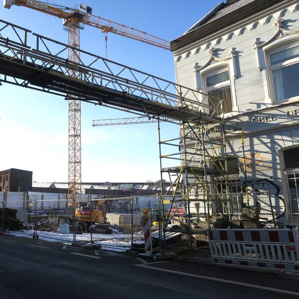 An einer Straße ist eine große Baulücke. Ein Kran und Baugerüste stehen dort hinter einem Bauzaun.