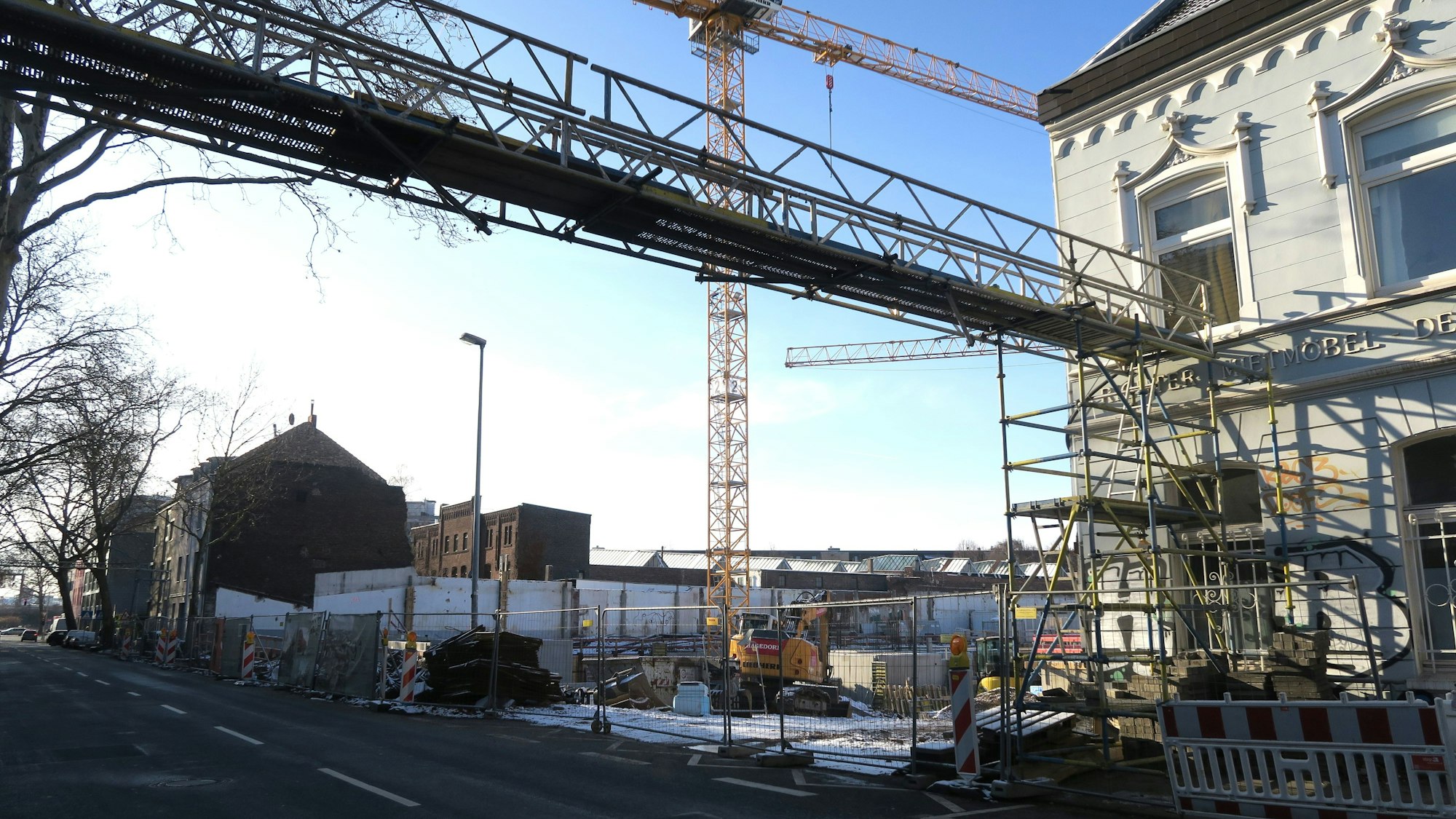 An einer Straße ist eine große Baulücke. Ein Kran und Baugerüste stehen dort hinter einem Bauzaun.