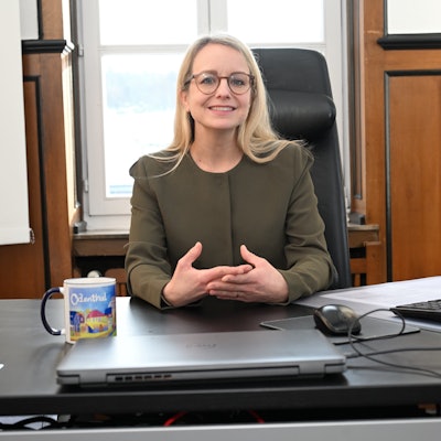 Eine Frau mit blondem, langen Haar und Brille sitzt an einem Schreibtisch.