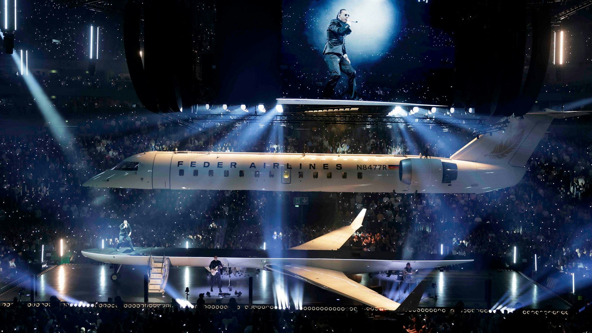 Apache 207 steht auf der Bühne der Lanxess-Arena. In der Mitte der Bühne steht ein Privatjet, der der Länge nach geteilt ist.