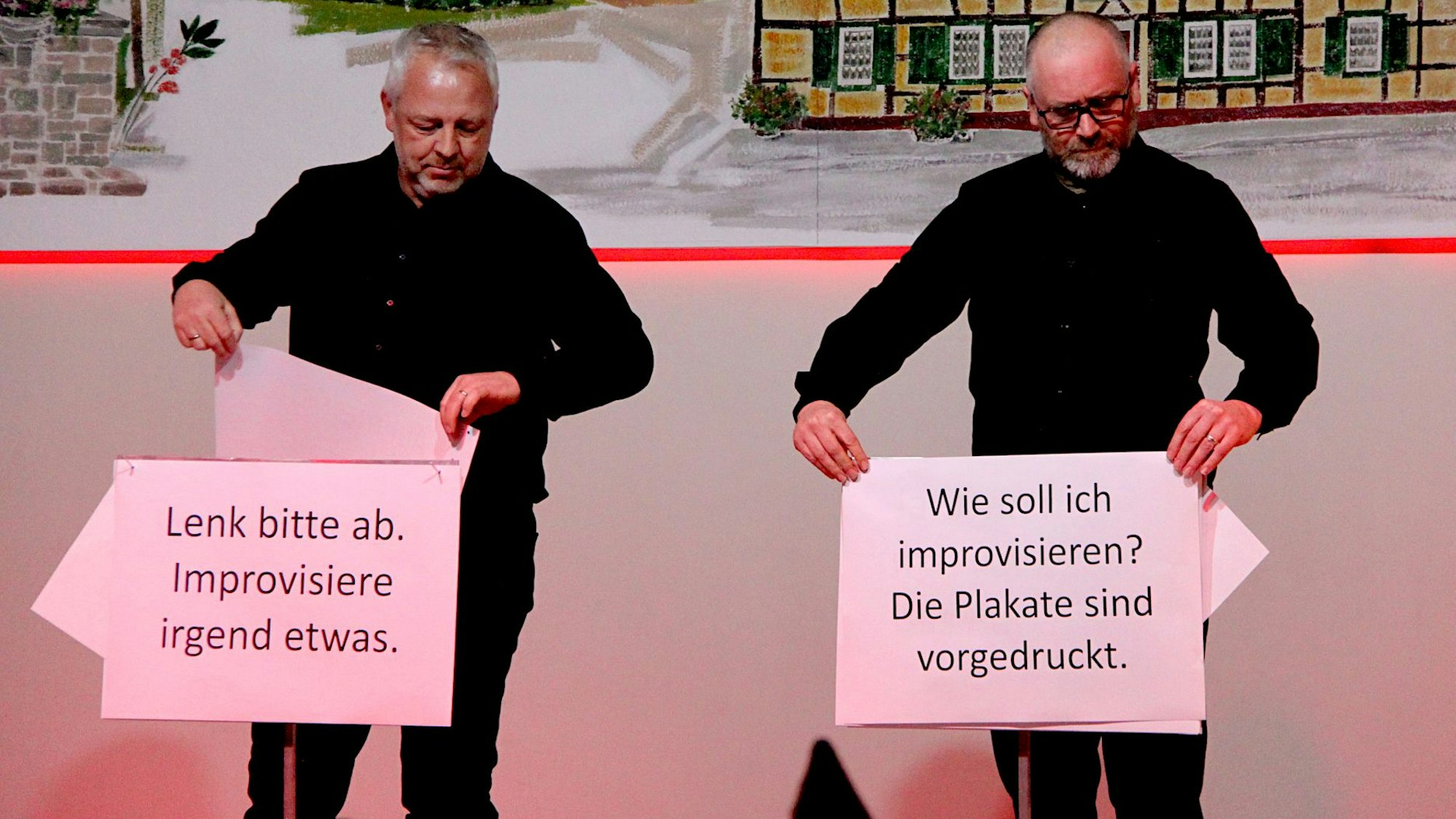 Zwei schwarz gekleidete Männer stehen auf der Bühne, sie halten mit Text beduckte große Papierseiten in den Händen.