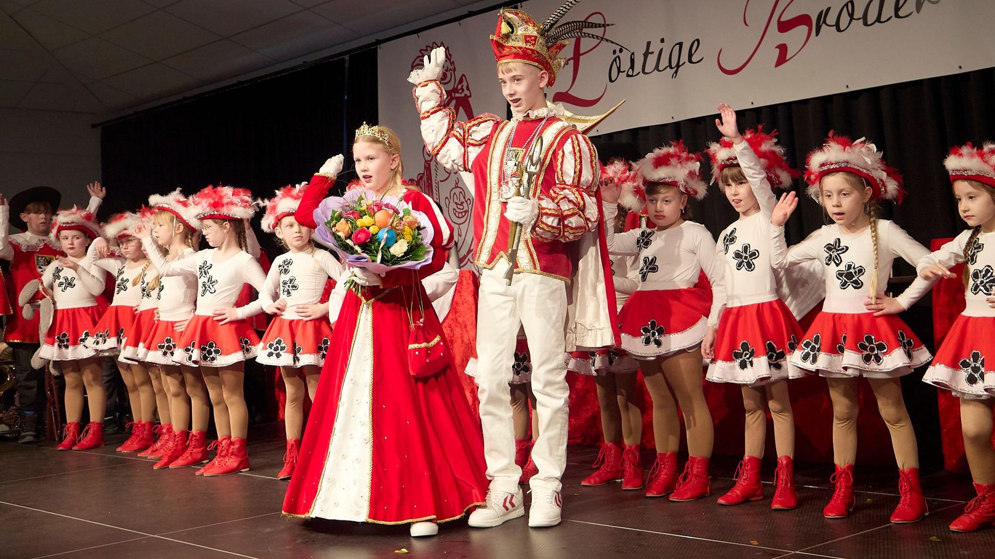 Auf der Bühne stehen Kinder der Tanzgarden der „Löstige Bröder“. Davor steht das Prinzenpaar und hebt die Arme zum Alaaf-Ruf in die Höhe.