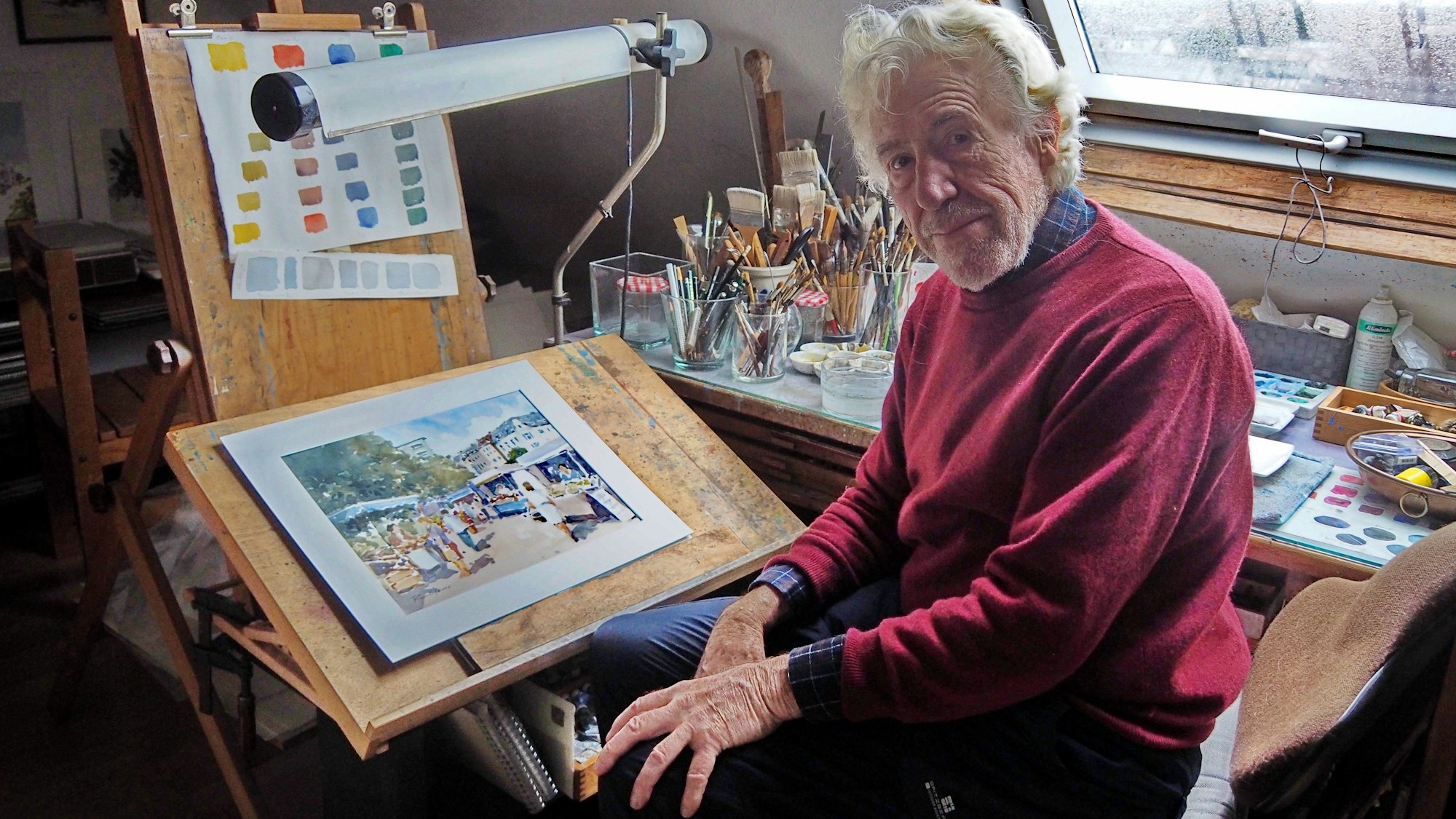 Ein älterer Mann sitzt an einem Maltisch unter einem Dachfenster.