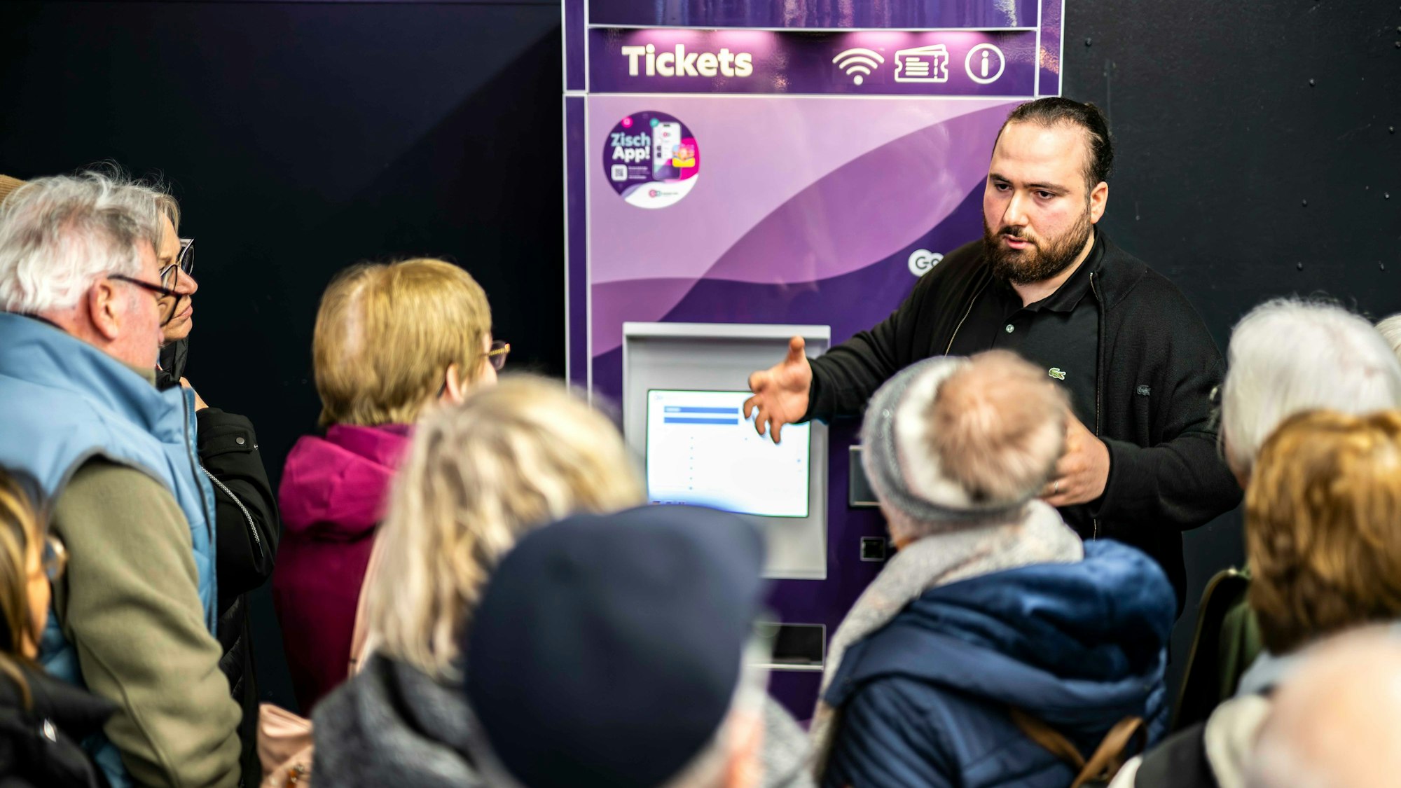 Ein Mann steht vor einem Ticketautomaten und gestikuliert. Ihm zugewandt stehen einige Leute.