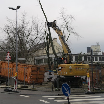 An einer Straßenecke ist ein Bauzaun, ein Lkw und ein Bagger sind dahinter zugange.
