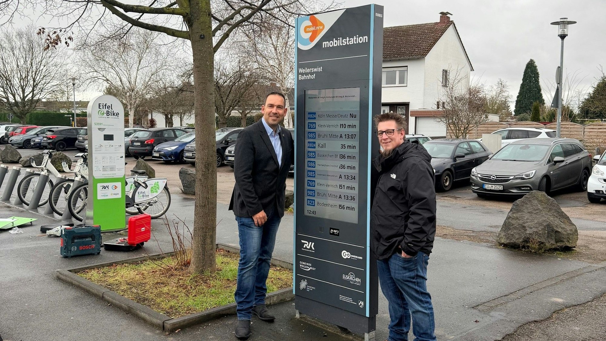 Dino Steuer (l.) und Christoph Bramer stehen an einer Mobilstation, sprich einer Anzeigetafel, auf der die Fahrtzeiten von öffentlichen Verkehrsmitteln abgebildet werden.