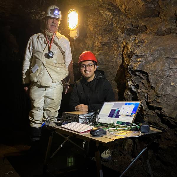 Das Bild zeigt einen Mann in Grubenmontur in einem Stollen, der neben einem Jungen steht, der an einem Tisch mit einem Computer sitzt.