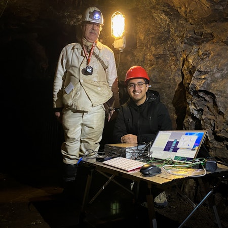 Das Bild zeigt einen Mann in Grubenmontur in einem Stollen, der neben einem Jungen steht, der an einem Tisch mit einem Computer sitzt.