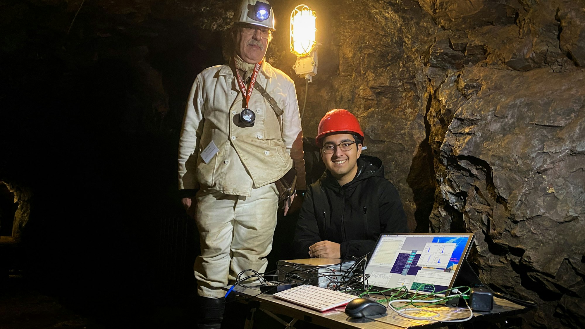 Das Bild zeigt einen Mann in Grubenmontur in einem Stollen, der neben einem Jungen steht, der an einem Tisch mit einem Computer sitzt.