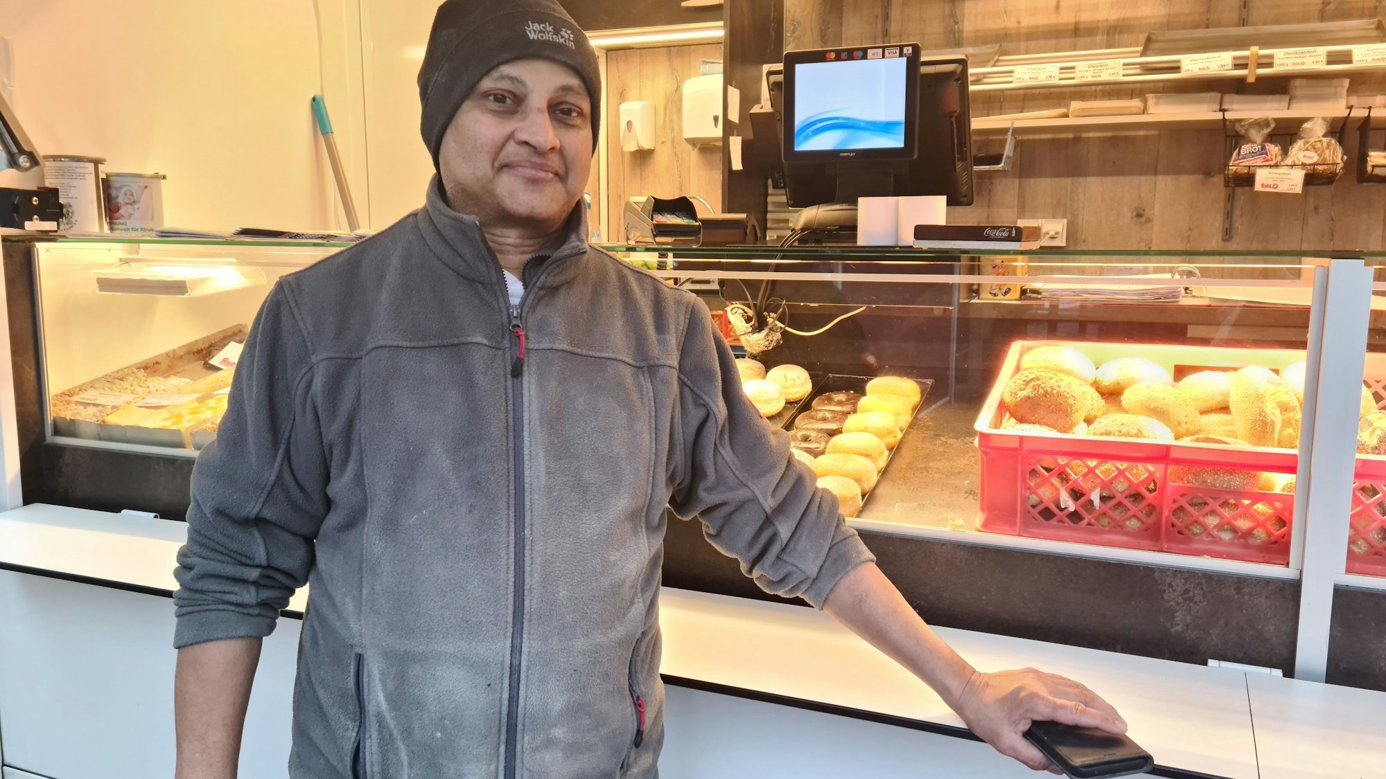 Ein Mann mit dunkler Haut, grauer Jacke und grauer Mütze steht vor einem Marktwagen, in dem Backwaren liegen.