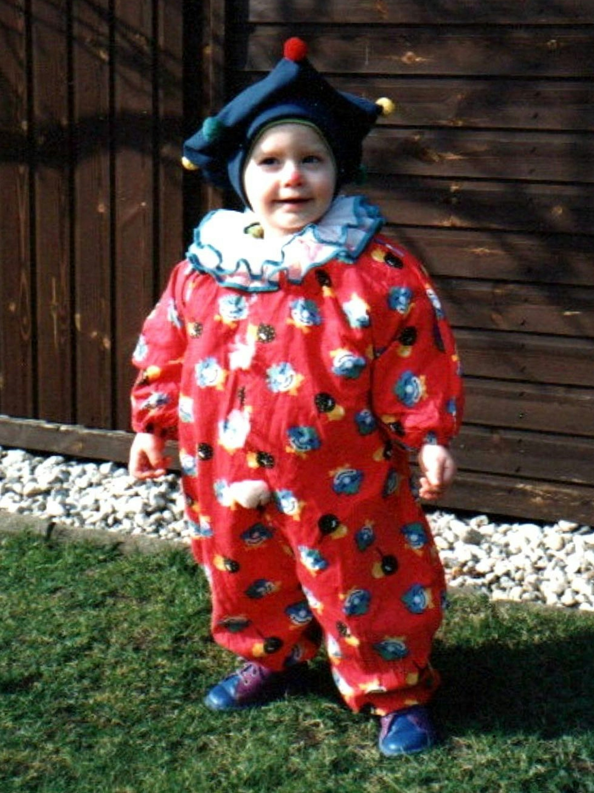 Marice Jakobs steht als Kind im Harlekinskostüm auf einem Rasenstück vor einem Holzhaus.